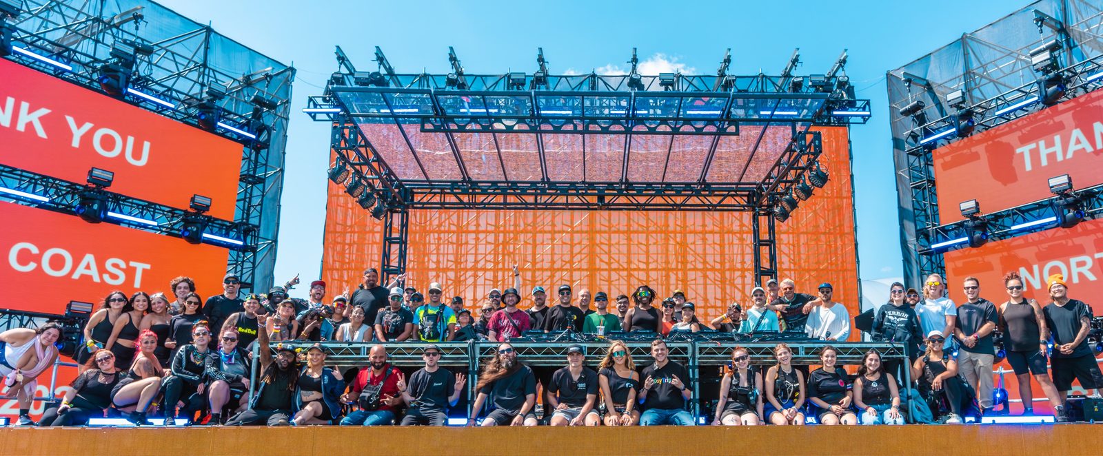 A festival crew of several dozen people gathered on a stage with an orange scrim, flanked by screens reading thank you North Coast