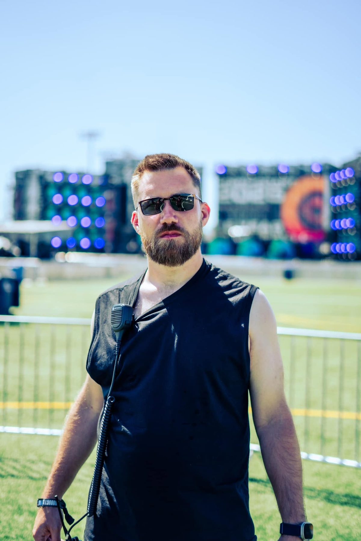 Sam standing on a festival field in a black shirt and sunglasses, radio mic clipped at his collar, stage screens behind him